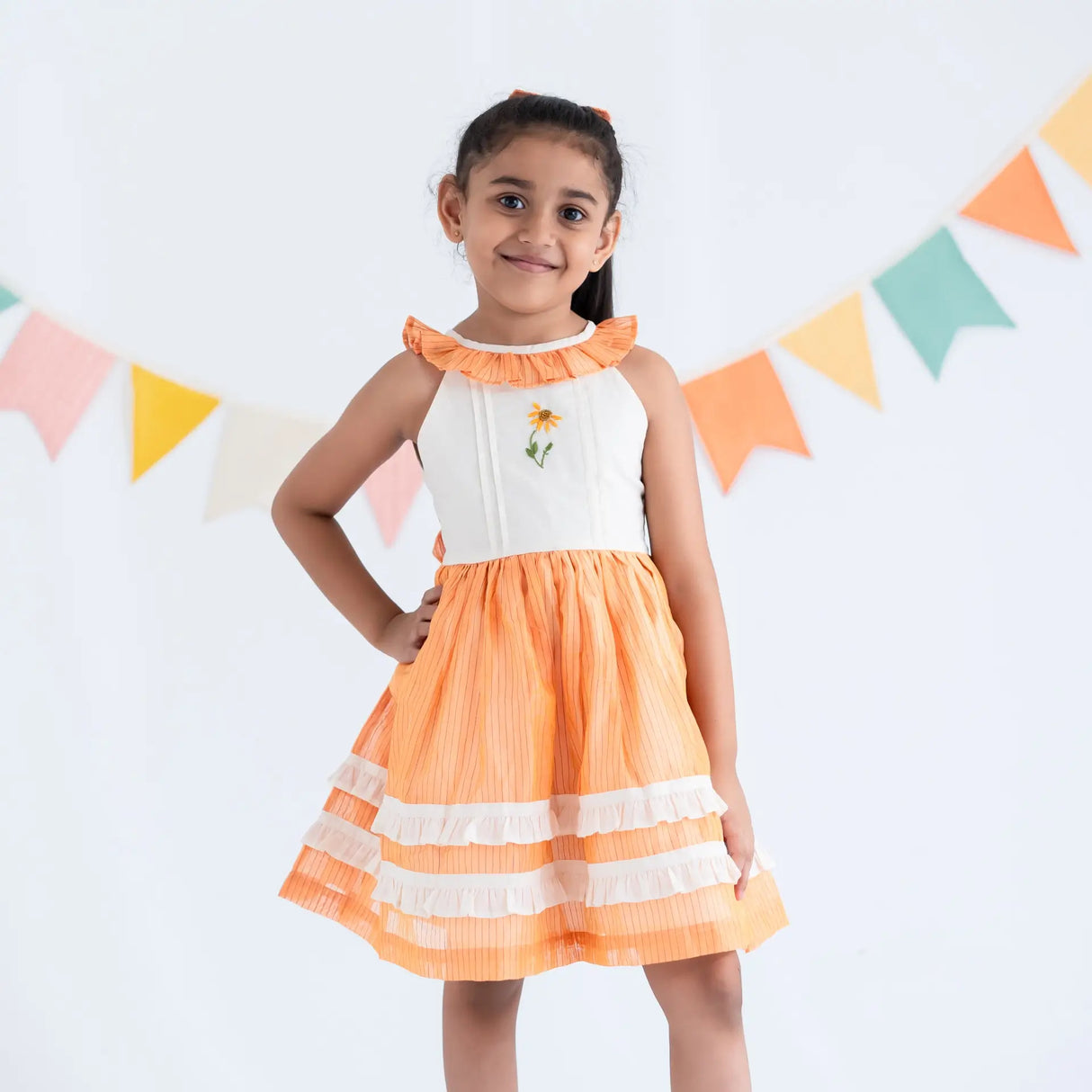A girl dressed in an orange hand embroidered zari striped chanderi sleeveless dress, standing with her hand on her hip.