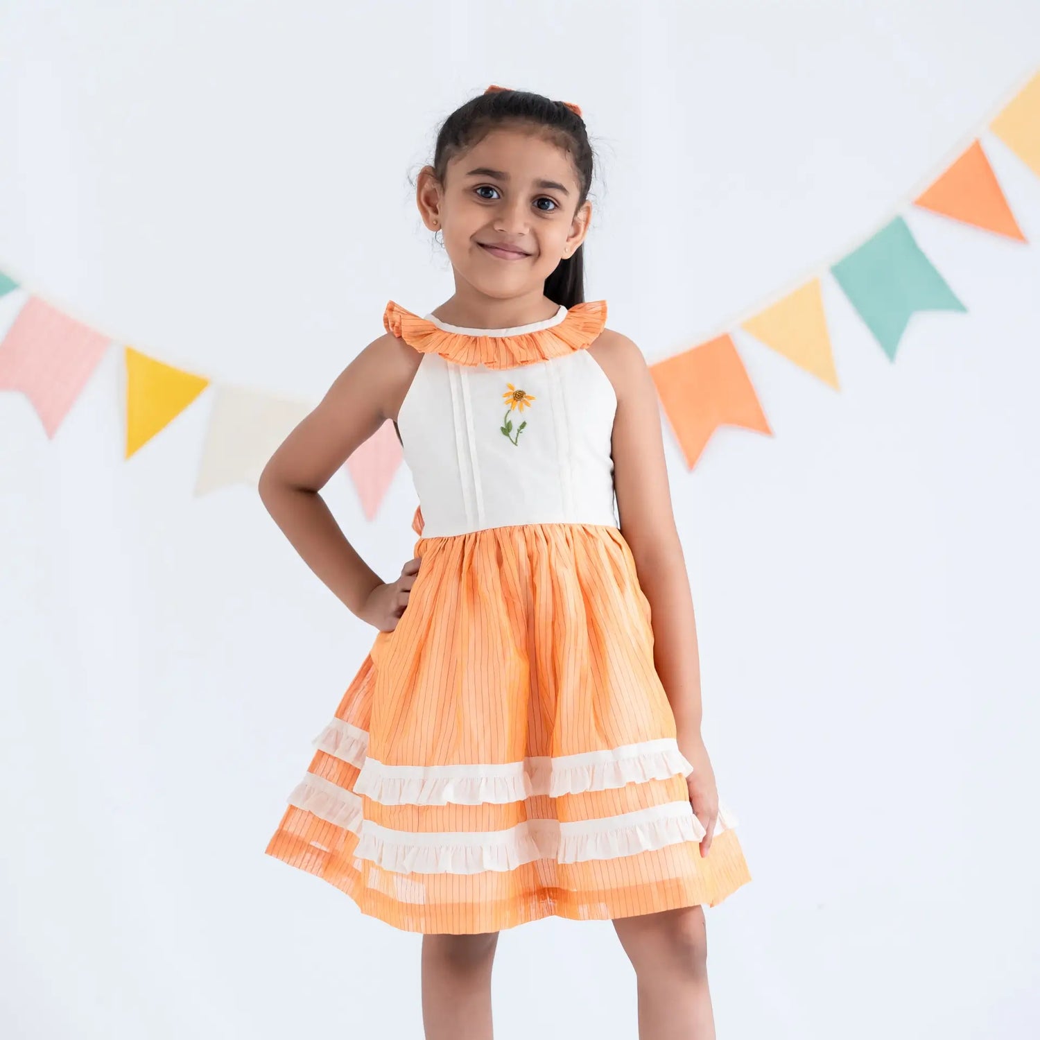 A girl dressed in an orange hand embroidered zari striped chanderi sleeveless dress, standing with her hand on her hip.