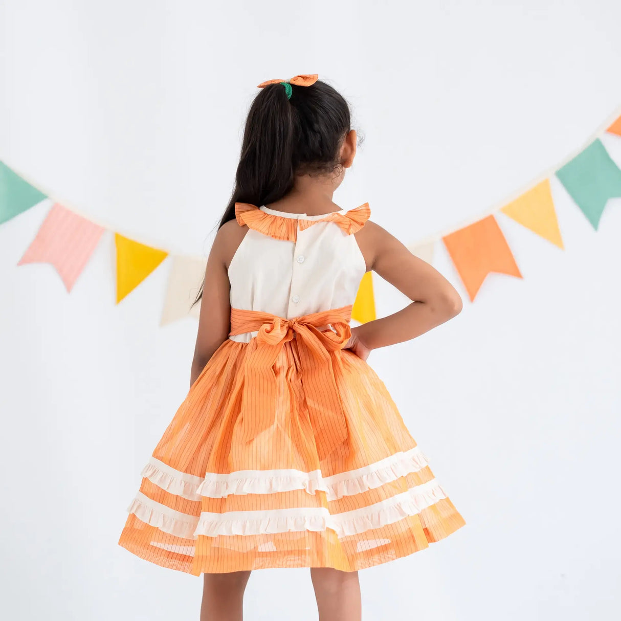 Back view of a girl dressed in an orange hand embroidered zari striped chanderi sleeveless dress.