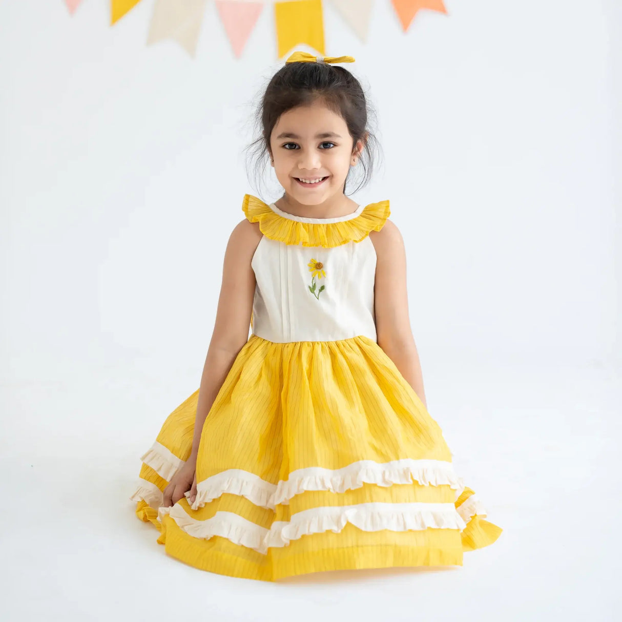 "A little girl seated, wearing a yellow hand embroidered zari striped chanderi sleeveless dress."