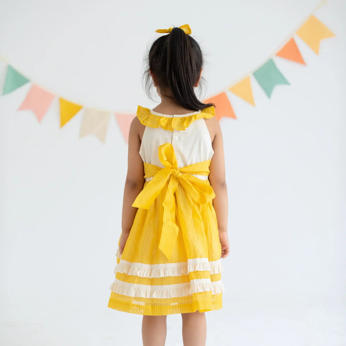 "Back view of a girl dressed in a yellow hand embroidered zari striped chanderi sleeveless dress."