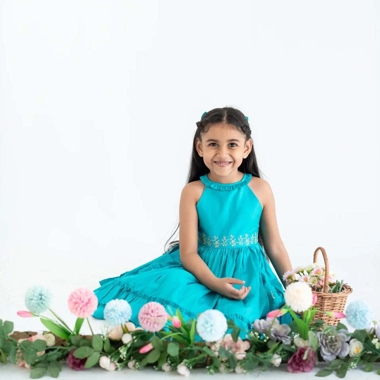 "A girl seated in a teal embroidered cotton satin sleeveless dress with pockets."