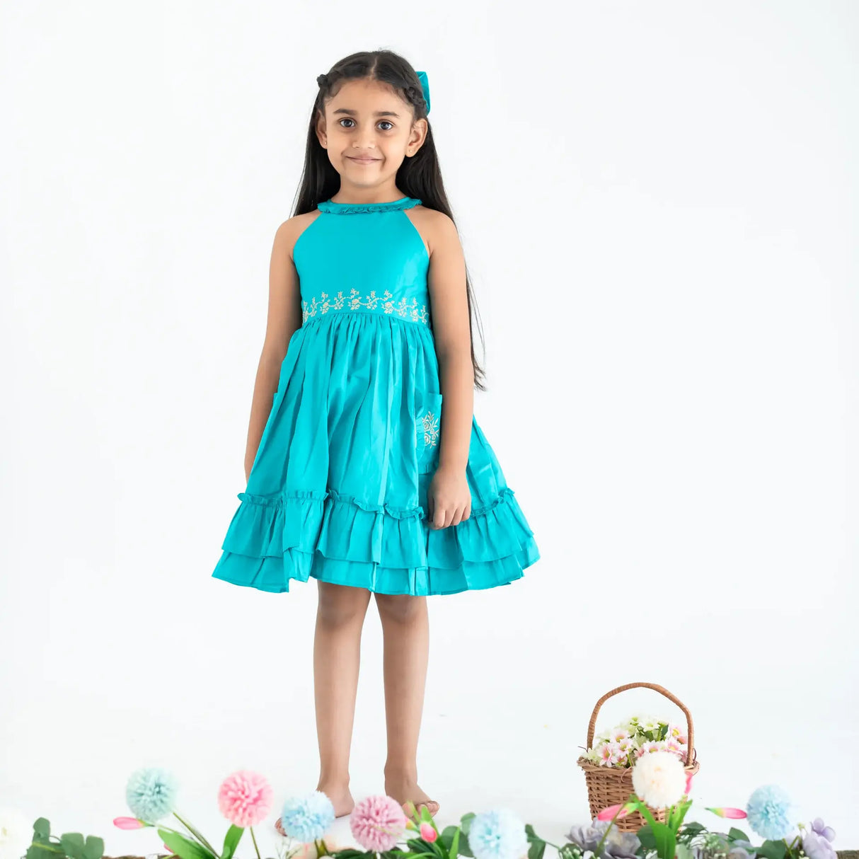 "A girl dressed in a teal embroidered cotton satin sleeveless dress with pockets."