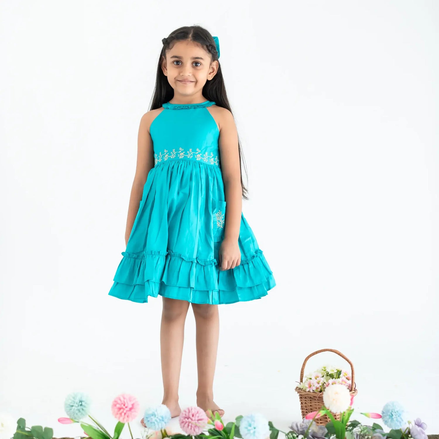 "A girl dressed in a teal embroidered cotton satin sleeveless dress with pockets."