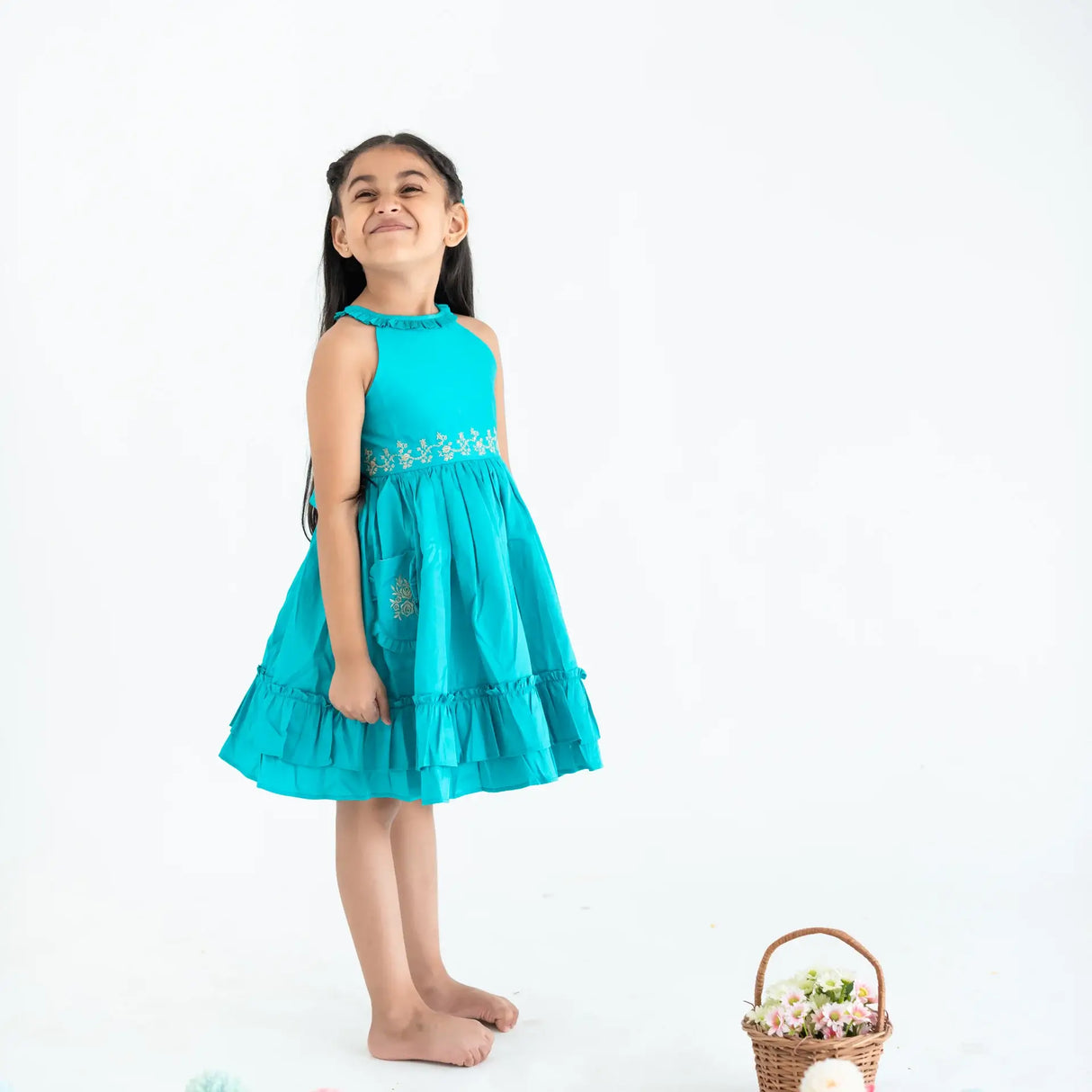 "Side pose of a girl dressed in a teal embroidered cotton satin sleeveless dress with pockets."