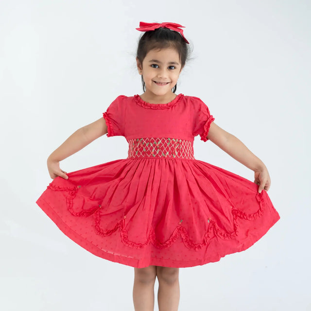 "A girl dressed in a coral swissdot hand smocked and embroidered cotton puffed sleeves dress, stretching the dress with her hands."
