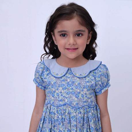 Close up image of a little girl in blue floral print dress with scalloped waistband, hand embroidered collar, puff sleeves and ruffled hem