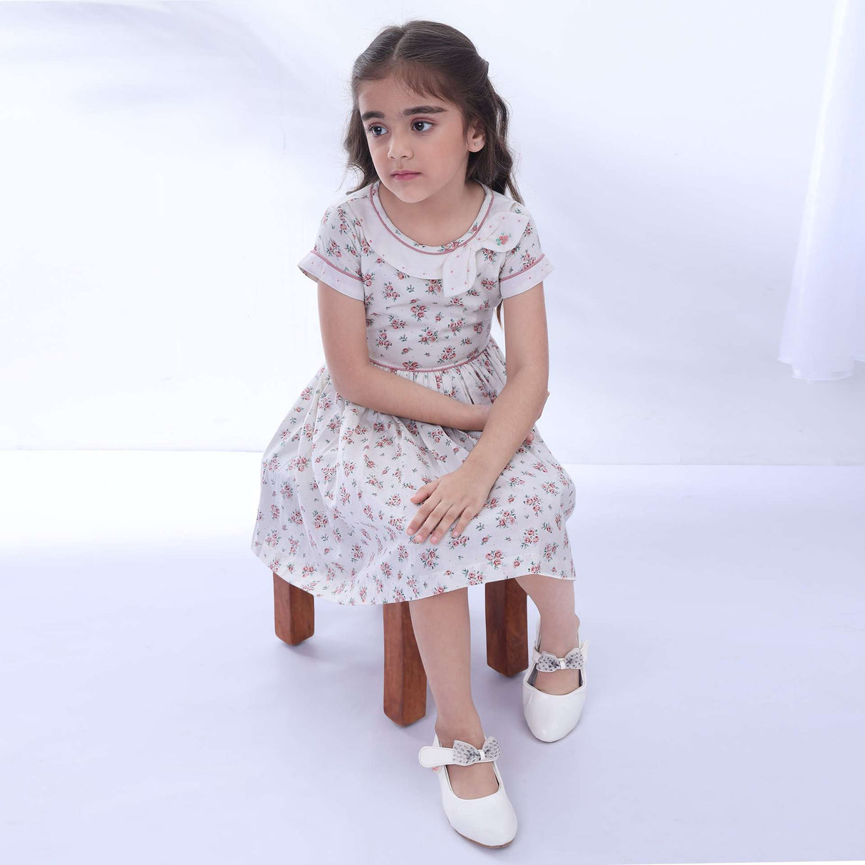 A seated little girl wearing ivory poplin floral-spray printed dress adorned with hand embroidery