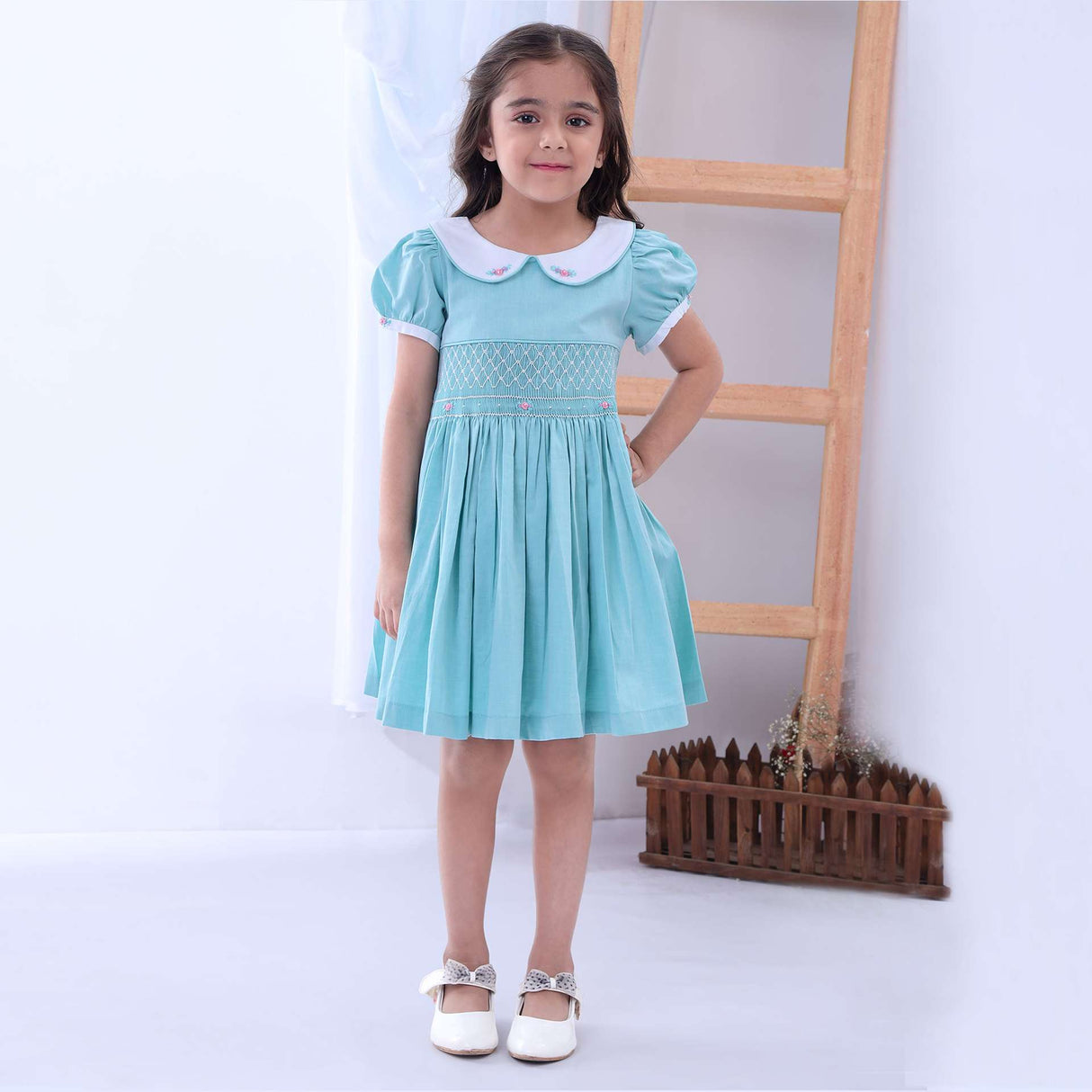 Girl poses in front of a ladder in a turquoise chambray classic smocked dress with embroidered collar