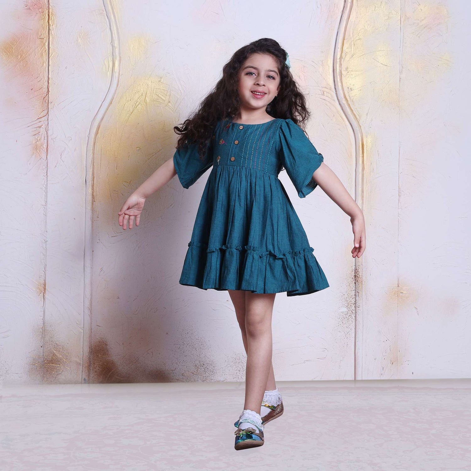 A little girl in peacock blue sparkler dress with hand embroidered details on bodice, ruffle hem, and gathered sleeves with button bridge