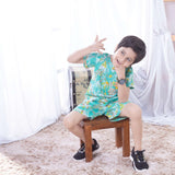 A little boy seated on a stool posing in green tropical palm printed cotton coordinated shirt and shorts set