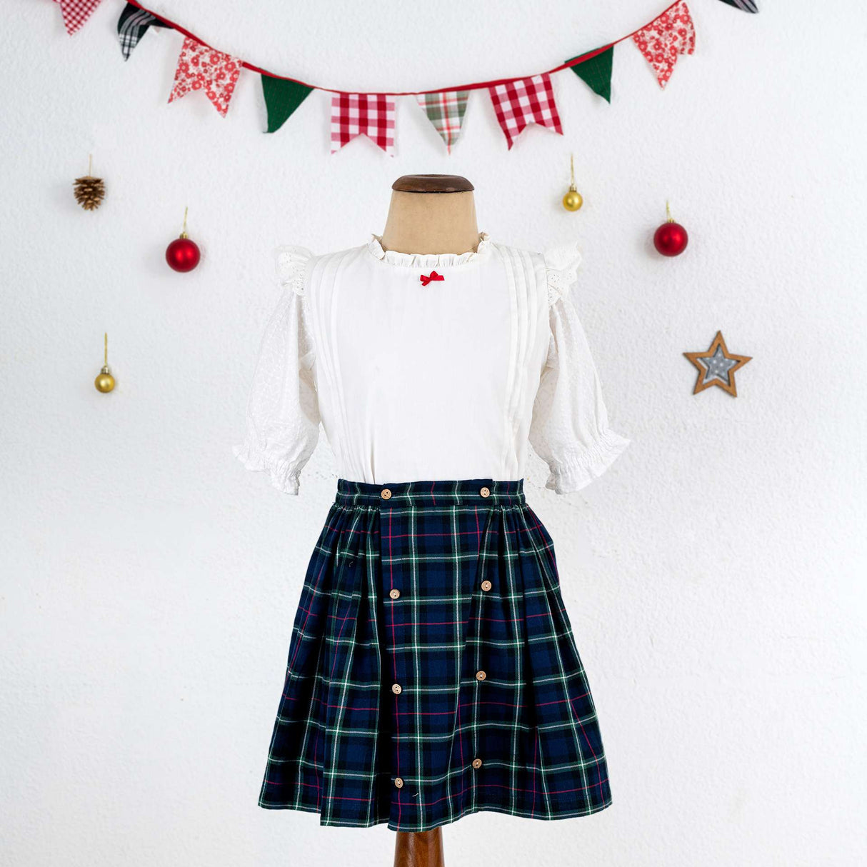 Mannequin shot of navy tartan wrap skirt paired with a frilly white cotton blouse with a little red satin bow