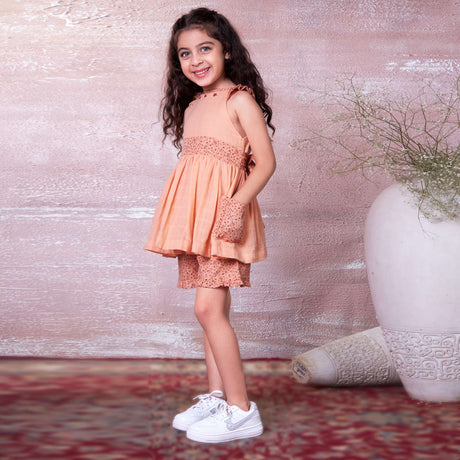 A little girl in peach gauze tunic with ruffles, hand embroidery and ties in a floral trim, with matching floral shorts