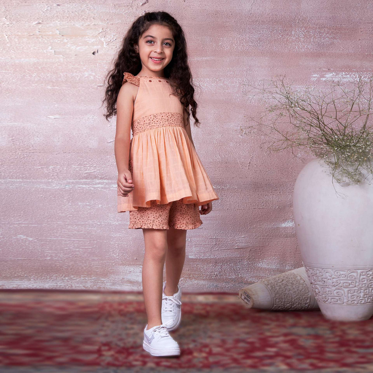 A little girl in peach gauze tunic with ruffles, hand embroidery and ties in a floral trim, with matching floral shorts