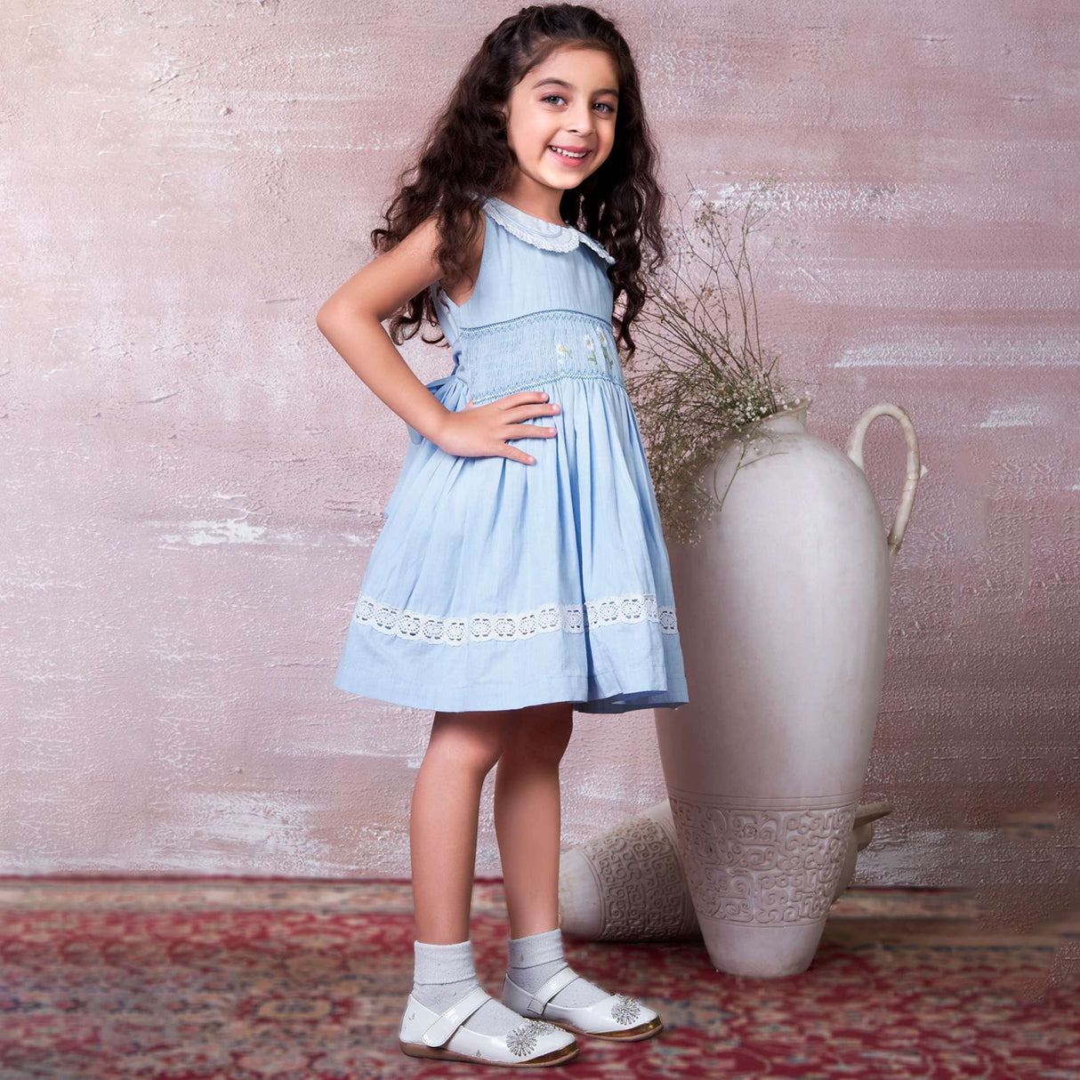 A little girl in blue hand-smocked dress with a lace insert on the skirt and a detailed collar with lace and cord piping