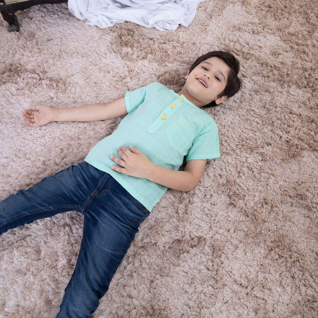 A boy laying down wearing aqua gauze boy's shirt with hand embroidered front pocket and yellow buttons