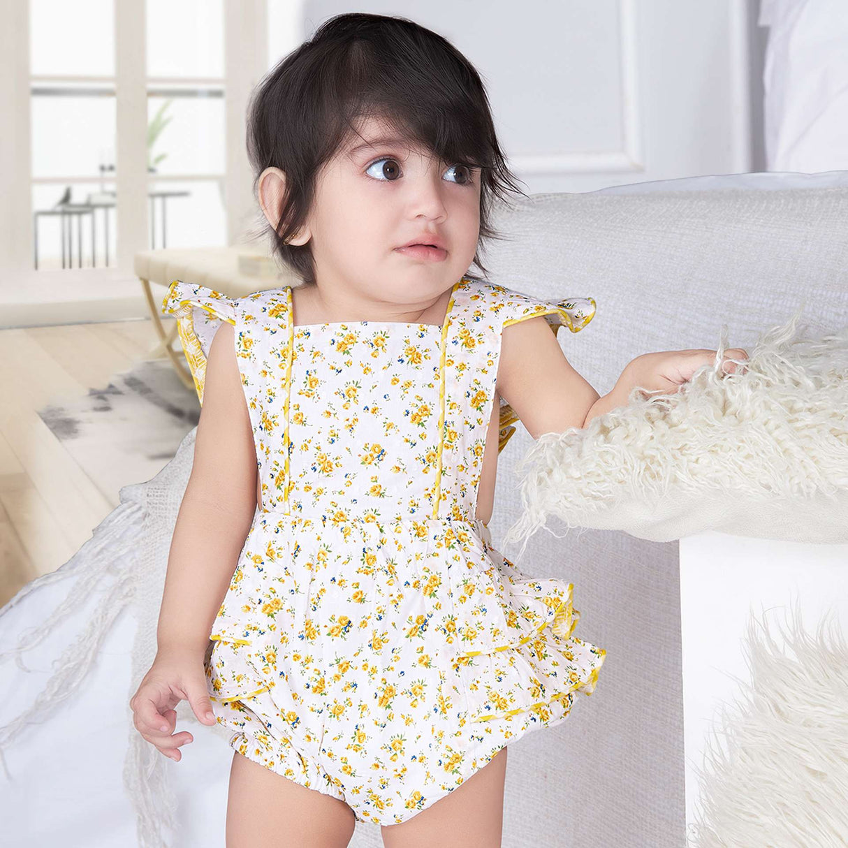 A baby girl in yellow floral printed onesie with back crossover straps and ruffles on the bottom with contrast trims