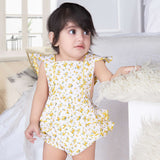 A baby girl in yellow floral printed onesie with back crossover straps and ruffles on the bottom with contrast trims