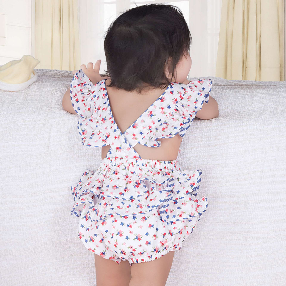 Back side of a baby girl in blue floral printed onesie with back crossover straps and ruffles on the bottom