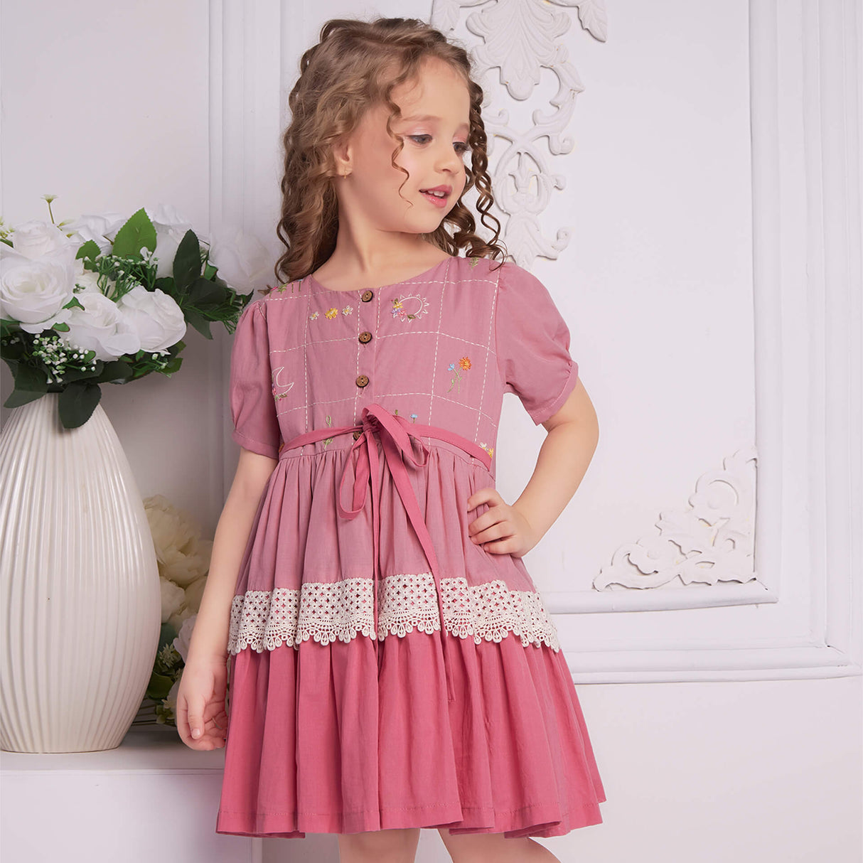 A girl in pink embroidered ombre dress with lace detail, tie up sash, and wood button fastening