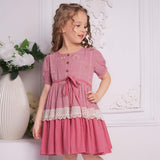 A girl in pink embroidered ombre dress with lace detail, tie up sash, and wood button fastening