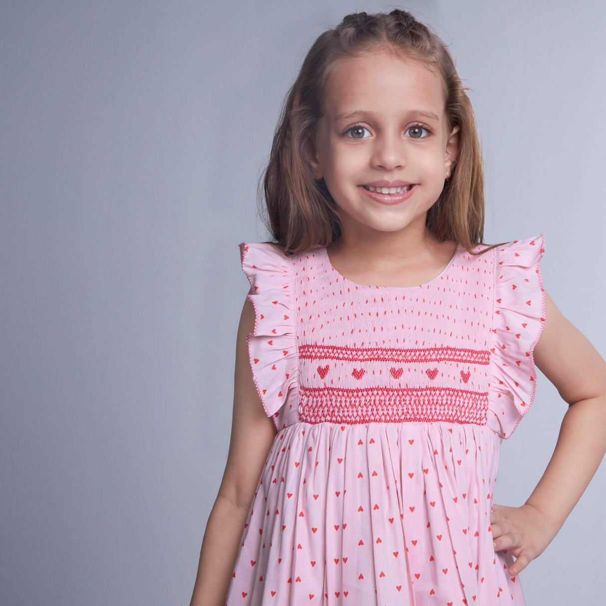 A little girl in a pink cotton dress with heart embroidery and smock, posing with a smile