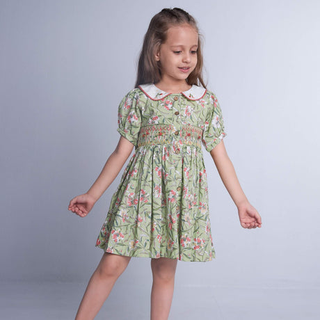 In a green floral smock dress, a child stands with hands straight, the collar adorned with delicate hand-embroidered details