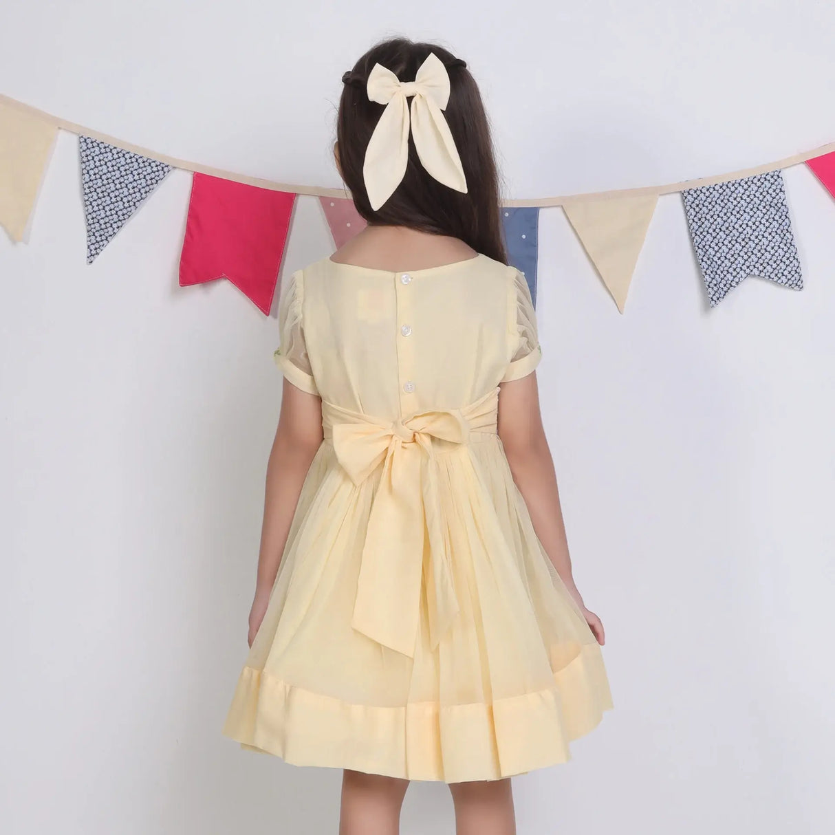 "Back view of a girl in a yellow hand-embroidered tulle dress, fastened with buttons and ties."