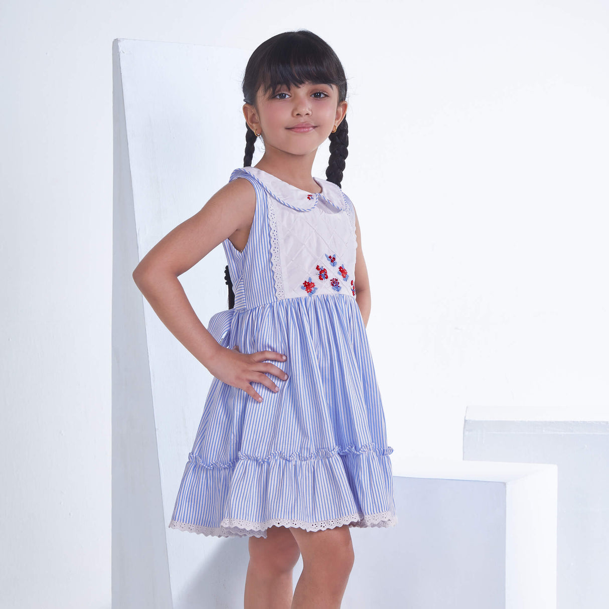 Posing from the side, a little one wears a blue striped embroidered collared dress adorned with ruffled hem
