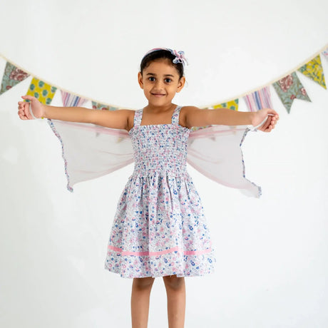 A girl dressed in a white floral printed strappy dress with attached wings at the back, playfully extending the wings.