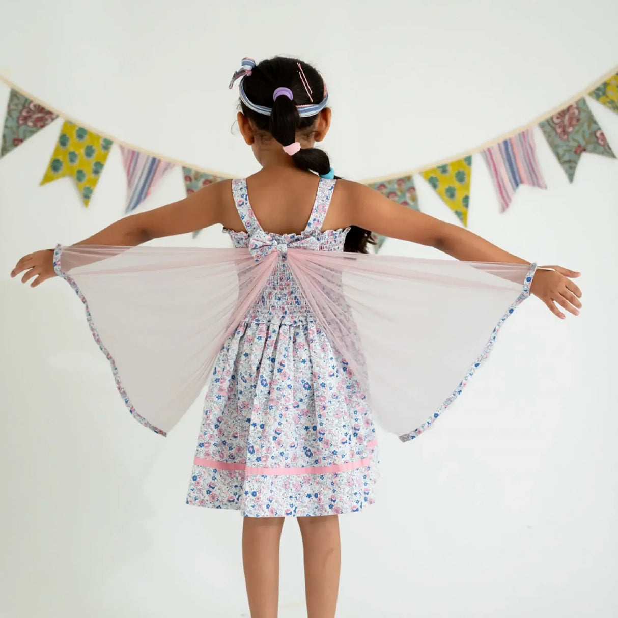 Back view of a girl dressed in a white floral printed strappy dress with attached wings at the back, playfully extending the wings.
