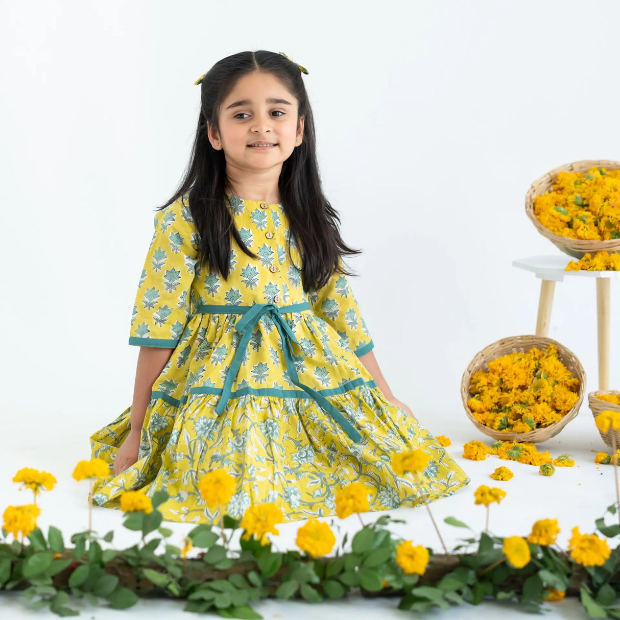 A girl seated, wearing a yellow and green printed long sleeves cotton dress with contrast trims.