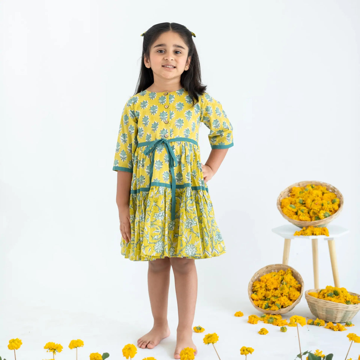 A girl dressed in a yellow and green printed long sleeves cotton dress with front opening buttons and ties.