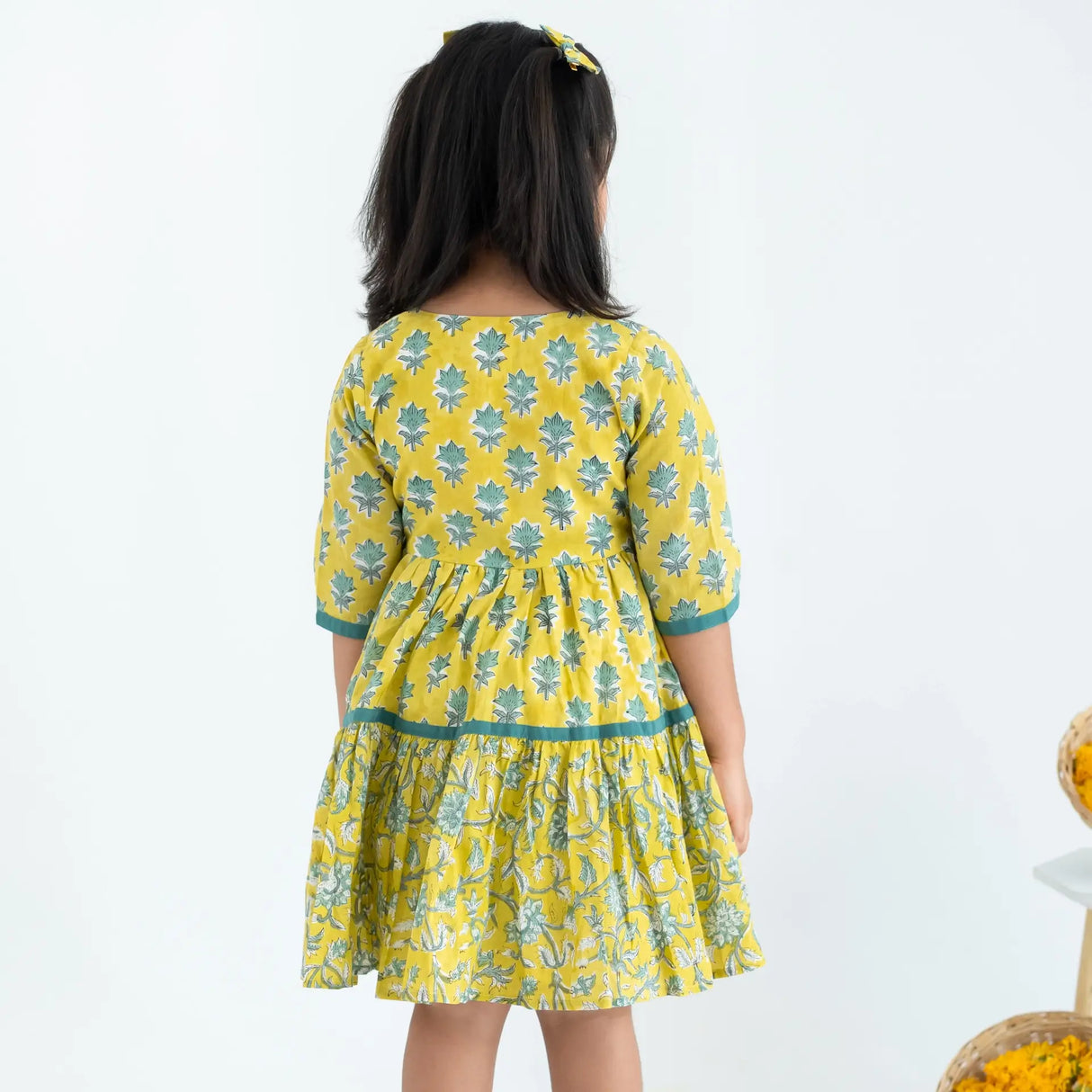 Back view of a girl dressed in a yellow and green printed long sleeves cotton dress.
