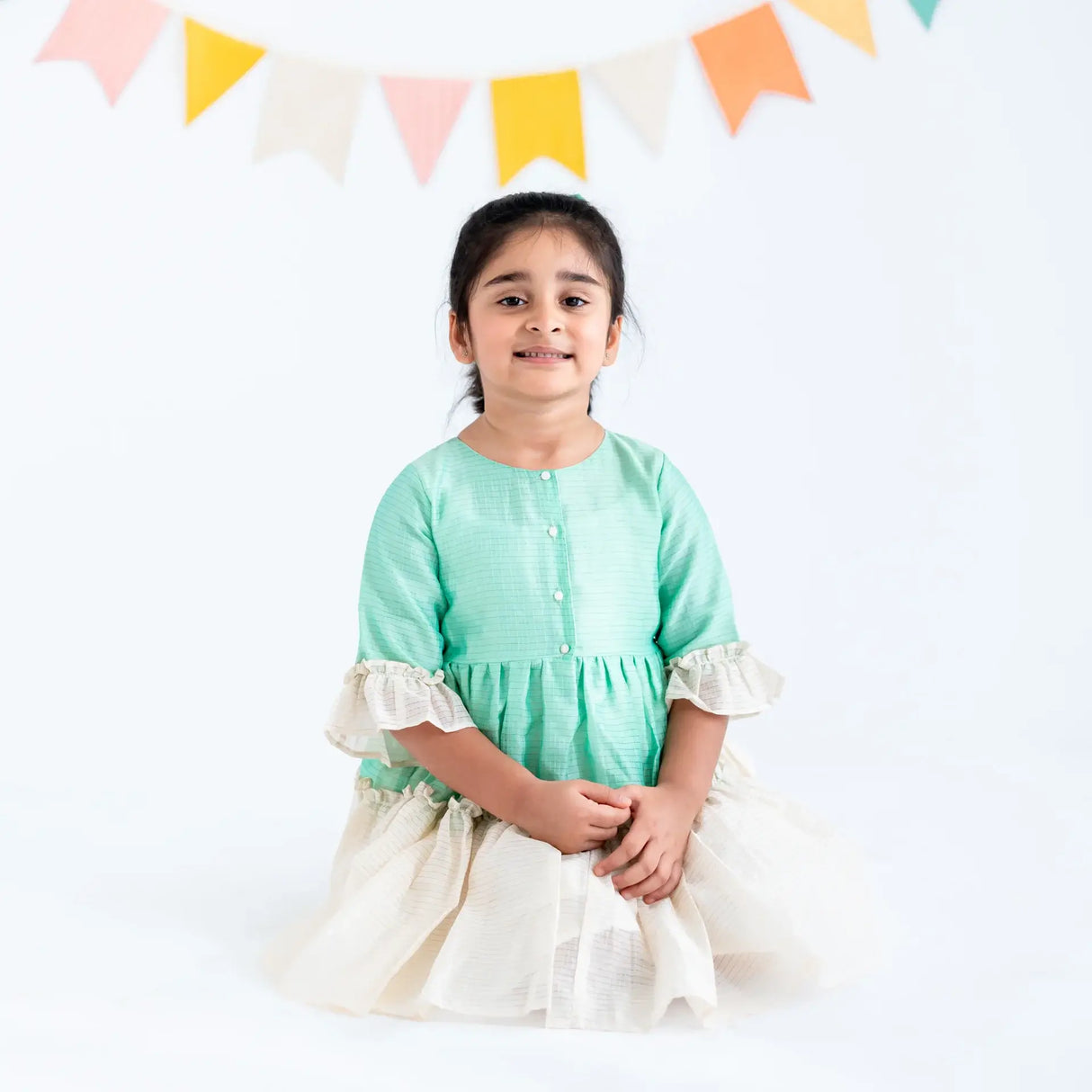 "A girl seated, wearing a green zari checked cotton silk half sleeves dress."