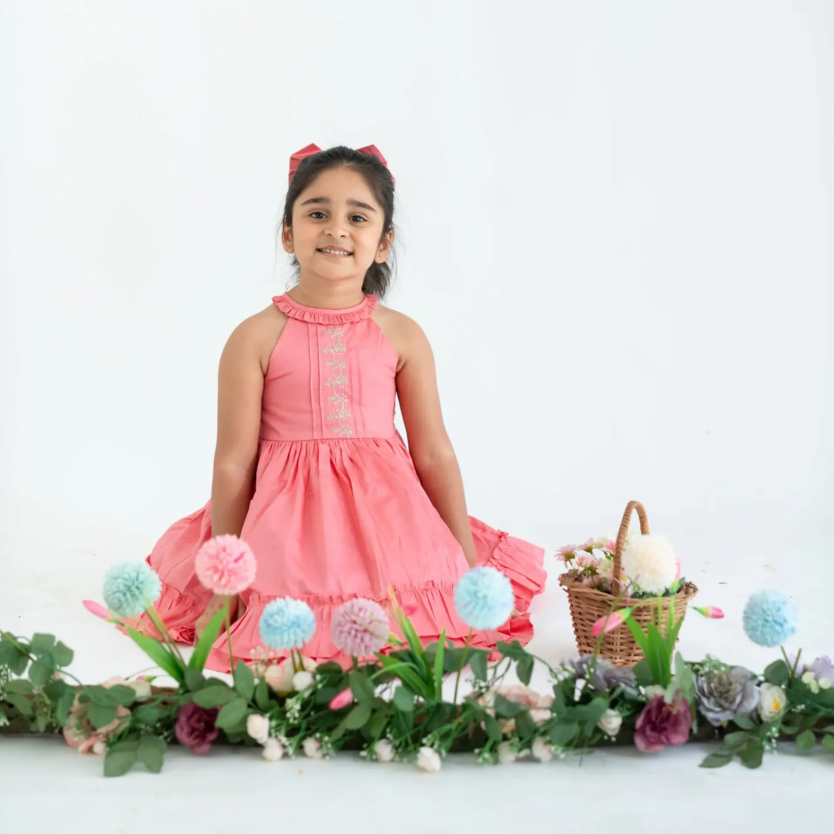 "A girl seated in a coral embroidered cotton satin sleeveless dress with pockets."