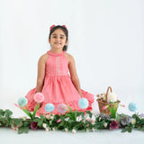 "A girl seated in a coral embroidered cotton satin sleeveless dress with pockets."