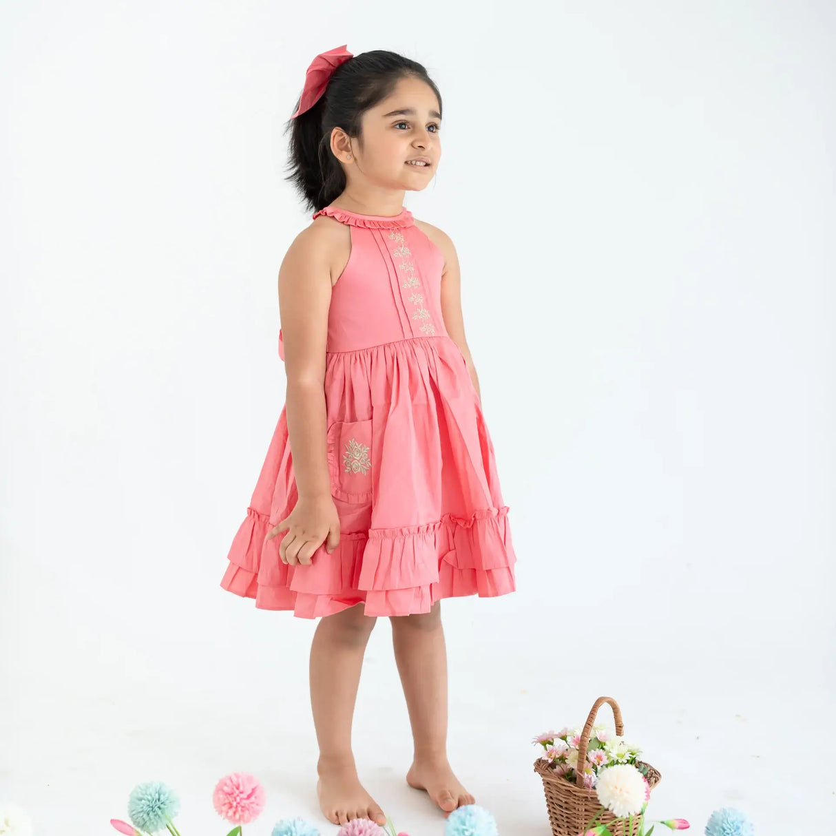 "Side pose of a girl dressed in a coral embroidered cotton satin sleeveless dress with pockets."