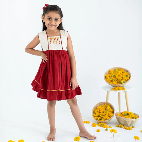 A girl dressed in a red sleeveless cotton dress with hand embroidery, standing with her hand on her hip.