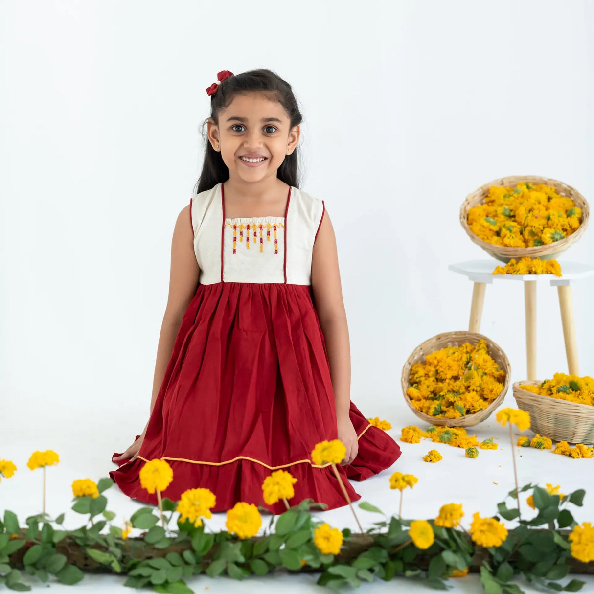 A girl seated, wearing a red sleeveless cotton dress with hand embroidery.