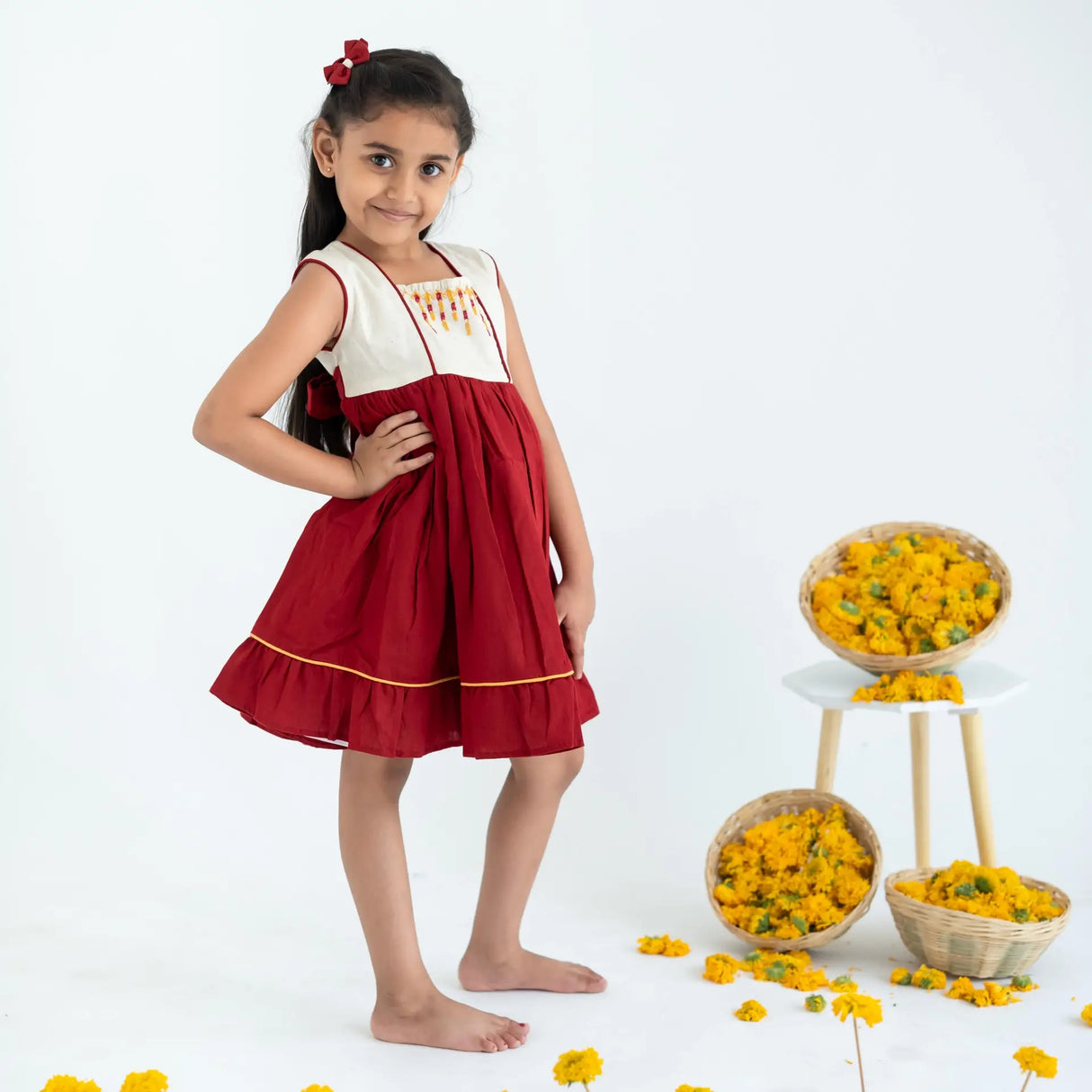 A girl in a red sleeveless cotton dress with hand embroidery, standing sideways with her hand on her hip.