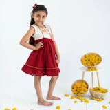 A girl in a red sleeveless cotton dress with hand embroidery, standing sideways with her hand on her hip.