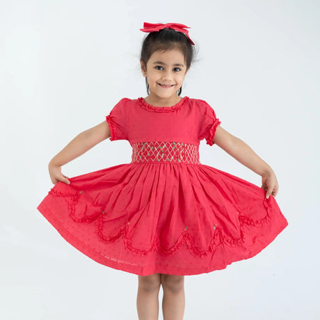 "A girl dressed in a coral swissdot hand smocked and embroidered cotton puffed sleeves dress, stretching the dress with her hands."