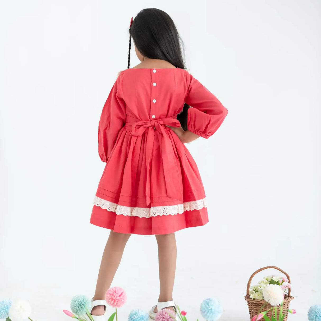 "Back pose of a girl dressed in a coral pleated yoke embroidered cotton long sleeves dress, fastened by ties and buttons."