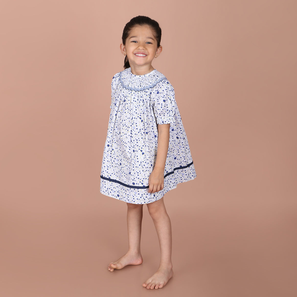 girl wearing the white and navy print hand-smocked dress with fabric trim along the hem