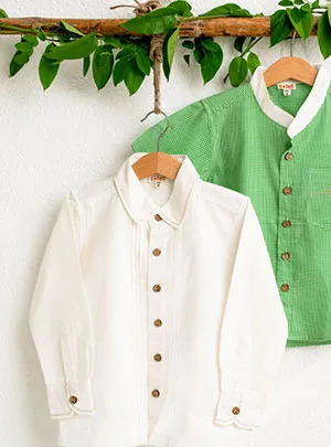 White lenin shirt and green checks boys' shirts, each with wood button fastenings and pockets, hang on hangers with leaves as prop