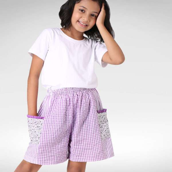A little girl in purple checked and striped patchwork shorts with lace trims on pockets and elastic waistband
