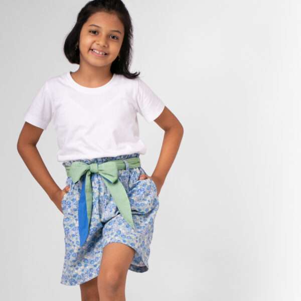 A young girl stands with a foot lifted wearing paperbag shorts in a blue floral with a green gingham belt