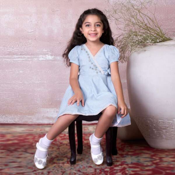 A little girl seated wearing blue striped cotton dress with delicate floral hand-embroidery and faux cross-over