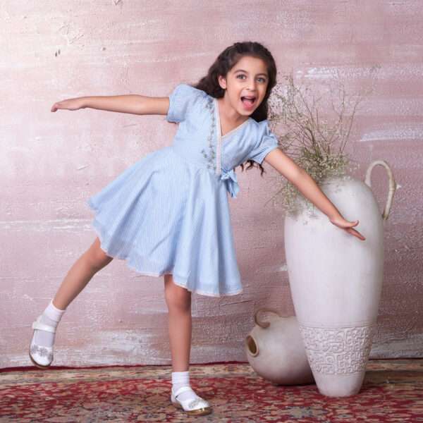 Little girl goofing around in striped blue striped muslin dress with crossover bodice, bow and hand embroidered details